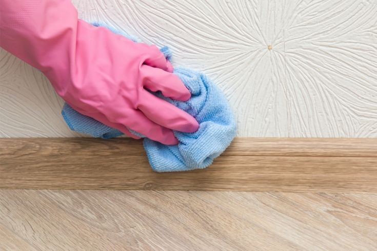 Hand in pink protective glove cleaning baseboard on the wooden floor from dust with rag at the wall. Early spring cleaning or regular clean up. Maid cleans house.